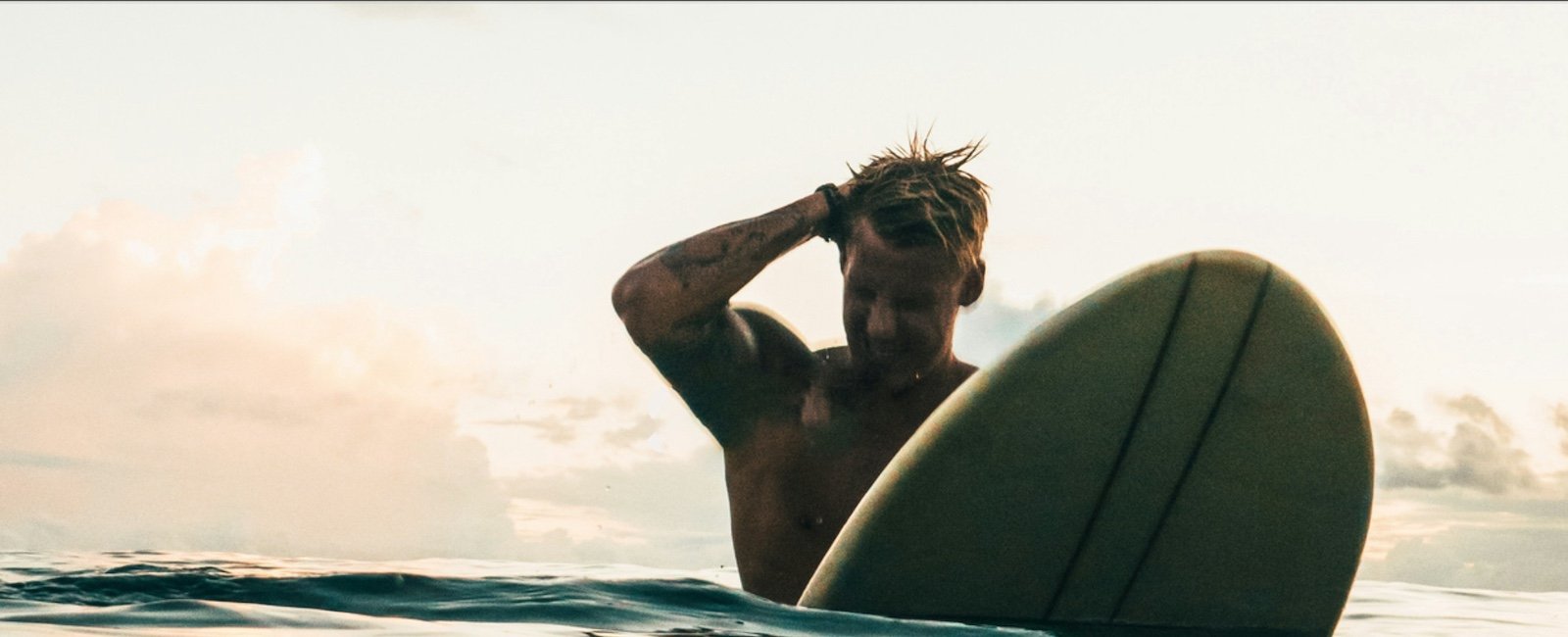 A surfer sitting on the surfboard with this right hand on is hair on a gold Sunset